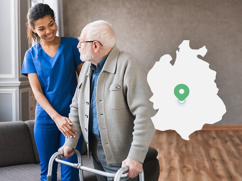 Einem älteren Mann wird zuhause von einer Pflegerin beim gehen geholfen.